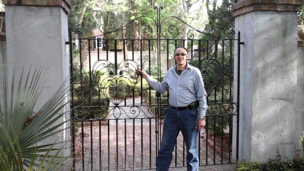 A man stands beside an ornate iron gate, partially opened, revealing a lush garden beyond. The scene suggests exploration or entry into a private space.