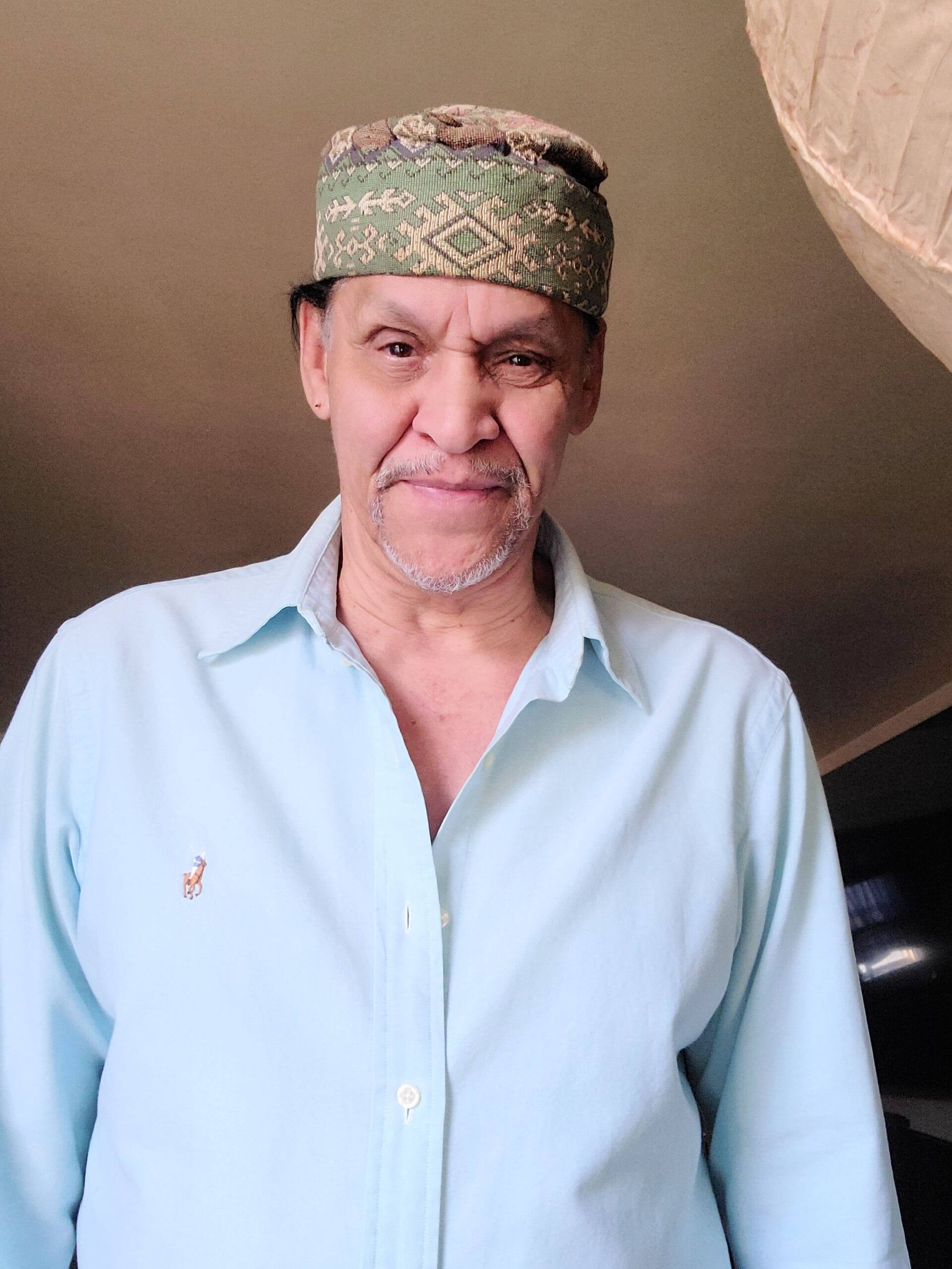 Man with a warm smile wears a light blue button-up shirt and a patterned green hat, standing in a comfortable indoor setting.